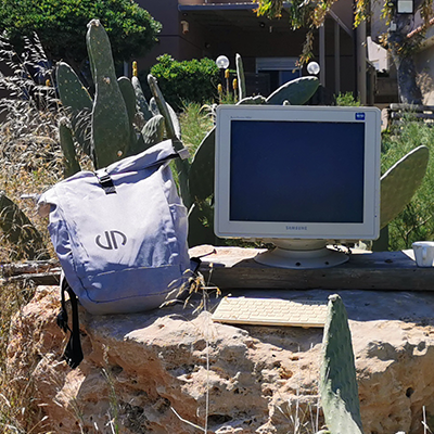Ein grauer Rucksack mit dem Logo von Lehleiter + Partner liegt neben einem alten Bildschirm inmitten von Felsen und Kakteen auf Kreta. Ein sonniges Urlaubsbild mit Remotework aus Griechenland.
