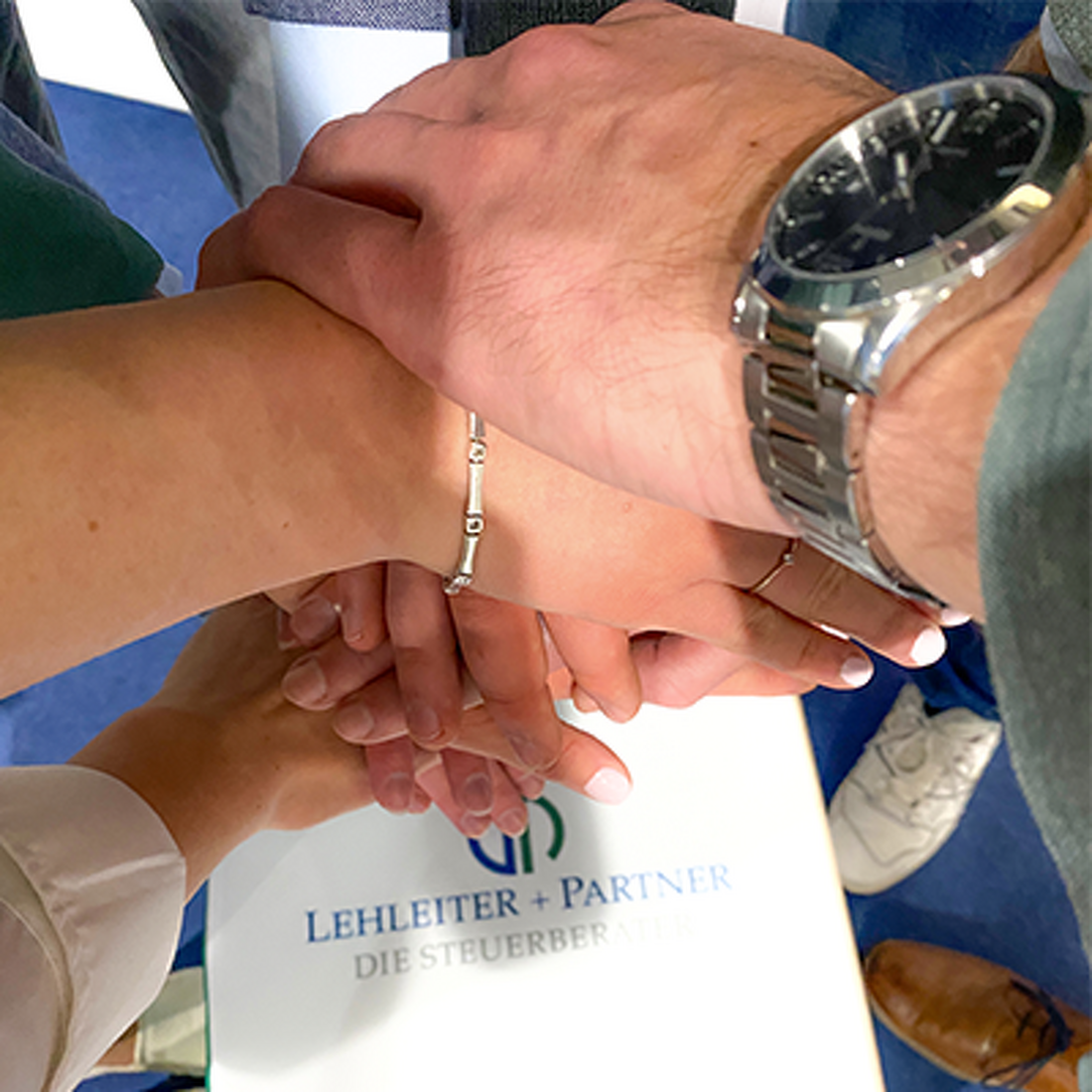 Steuerfachwirte, Steuerfachangestellte und Steuerberater von Lehleiter + Partner haben die Hände als Team übereinander gelegt. Darunter ist ein Sitzwürfel mit dem Logo von Lehleiter + Partner zu sehen.