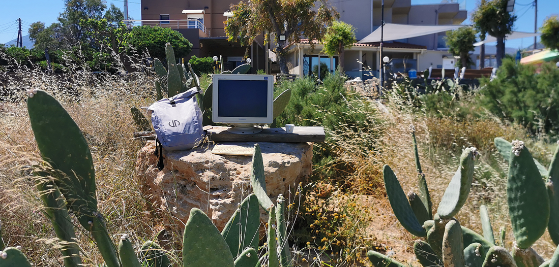 Ein grauer Rucksack mit dem Logo von Lehleiter + Partner liegt neben einem alten Bildschirm inmitten von Felsen und Kakteen auf Kreta. Ein sonniges Urlaubsbild mit Remotework aus Griechenland.