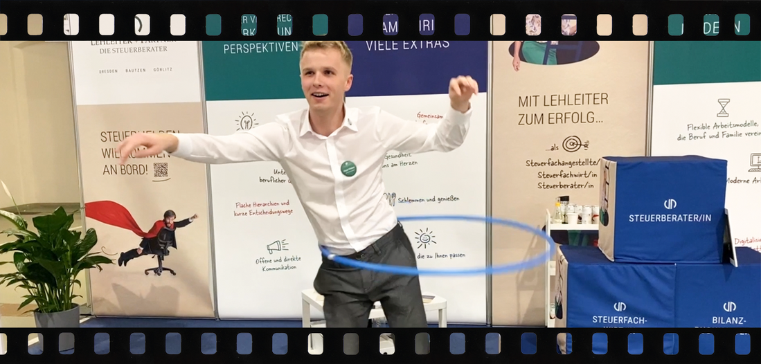 Ein Mitarbeiter von Lehleiter + Partner steht auf dem Messestand und versucht sich lächelnd am Hula Hupp. Im Hintergrund sind verschiedene Sitzwürfel zu sehen mit der Aufschrift Steuerberater, Steuerfachwirt und Steuerfachangestellte sowie die Messewände von Lehleiter + Partner.
