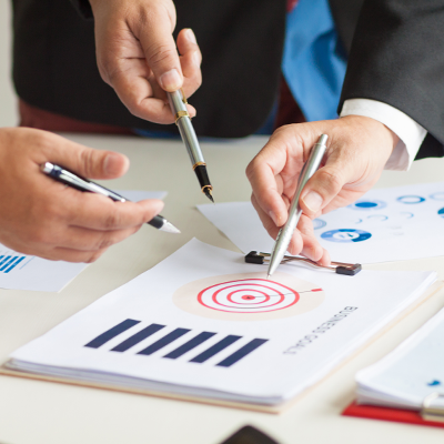 Businessbesprechung mit Businessplan und 3 Händen, welche mit Stiften auf den Businessplan zeigen.