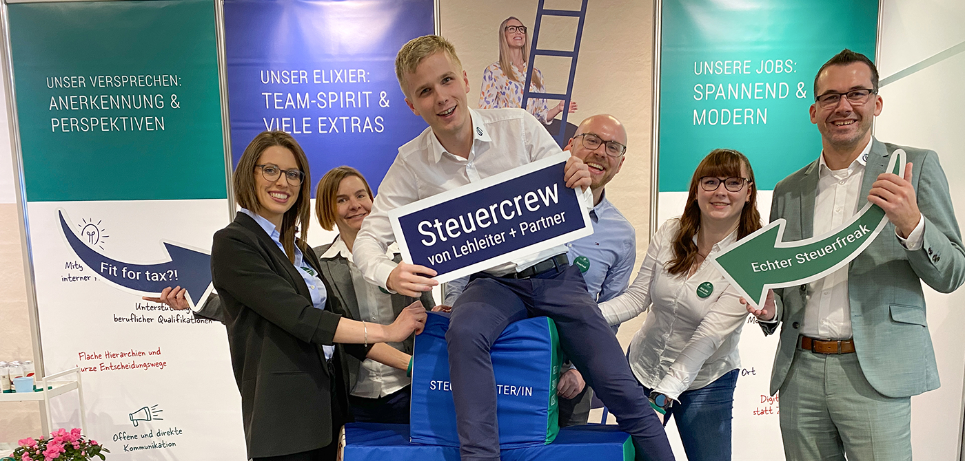 Sechs Mitarbeiter von Lehleiter + Partner stehen auf dem Messestand. Zu sehen sind Sitzwürfel mit der Aufschrift Steuerberater und Steuerfachwirt. Ein Mitarbeiter sitzt auf diesen und hält ein Schild in der Hand mit der Aufschrift: Steuercrew von Lehleiter + Partner. Alle lächeln und haben Spaß.